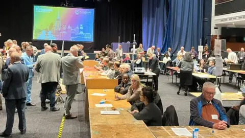 The scene at Portsmouth Guildhall during the election count. Lots of people are standing around while others are seated at long wooden tables. At the back of the hall is a black stage with blue curtains pulled to one side and a large screen showing the Portsmouth City Council logo in rainbow colours.