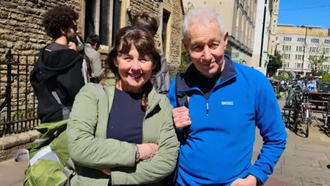 Eoin McCaul/LDRS A woman and man stand together and smile at the camera. The woman wears a green jacket, navy top and carries a green backpack. The man wears a blue fleece and carries a backpack on one shoulder. In the background are a number of buildings, people and parked bikes.