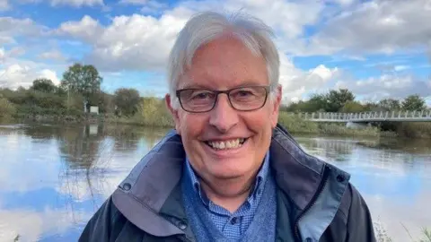 Glyn Marshall smiling. He is standing in front of the River Severn. There are trees and a bridge behind him. 