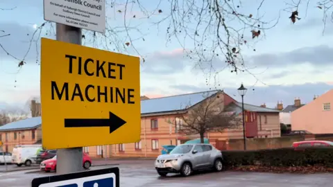 A car park with a yellow sign that reads ticket machine and a black arrow directing people toward it. Cars are parked in the car park. 