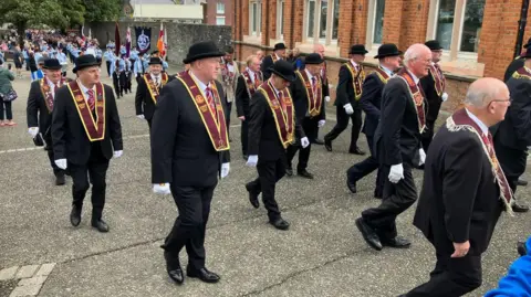BBC A group of men wearing black suits and black bowler hats marching in a parade.