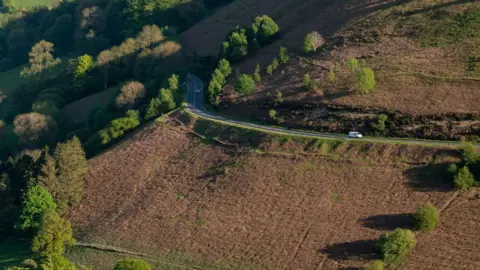 Getty Images A winding road can be seen going through rows of trees with a sharp drop off a mountain side. A van is driving along the road.