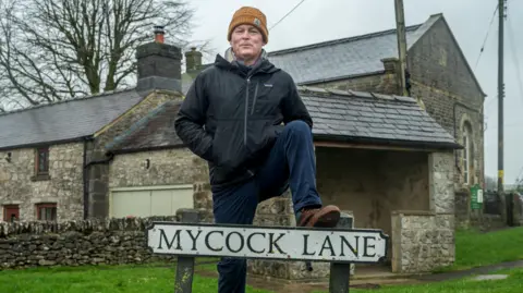 A smiling man in a raincoat and yellow hat stand with one foot resting on a road sign that says "Mycock Lane"