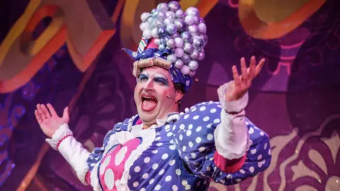 The Grove Theatre Dunstable Will Kenning, dressed up as a dame, wearing heavy make-up, a wig, including curlers and bubbles, and a blue spotted dress with big puffy sleeves. 