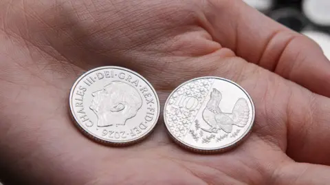 New 10p coin in the palm of someone's hand with one coin on the side showing an image of King Charles and dated 2026, with another coin on the reverse side showing a capercaillie.