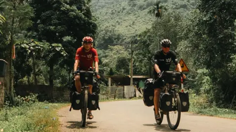 Kohler family Two men cycle towards the camera on a country road surrounded by foliage. Their cycles are laden with panniers. A green-covered mountain rises in the background. 