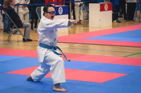 A teenage boy wearing a white karate outfit performs a punching move on a blue and red foam mat. He is wearing a blue karate belt, and glasses with a black frame. Behind him is a female judge sitting on a metal chair.