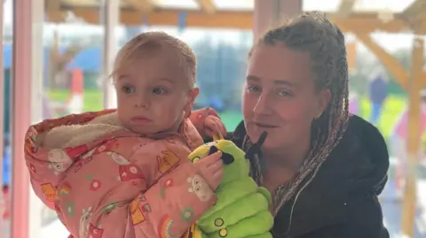 Becky Robinson holding her daughter who has a toy caterpillar. She has braided hair and is wearing a black coat. Her daughter has blonde hair and is wearing a Peppa Pig coat. In the background is a doorway leading to a garden.