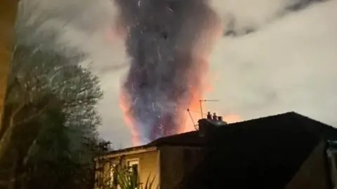 Smoke rising from behind a house during a fire at a paper mill in Fife. The smoke is grey and orange coloured against a dark, overcast background. The top of a house can be seen in the foreground alongside the outline of a tree.