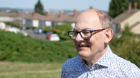Paul is wearing a white and blue patterned shirt with houses on. He is smiling and wearing glasses.