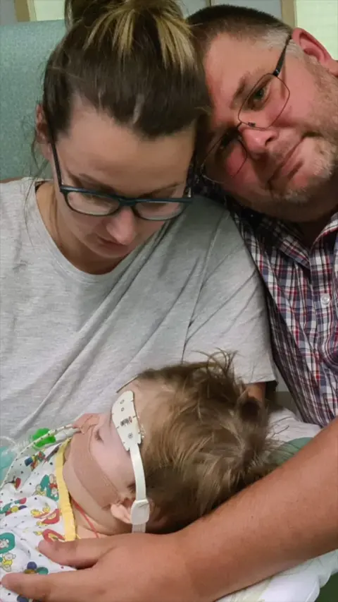 A man and a woman holding a toddler with a tube up their nose.