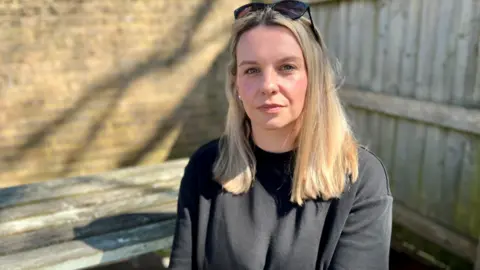 Rachel Moore looking at the camera has blonde hair and blue eyes, she has sunglasses on top of her head and is wearing a black t-shirt.