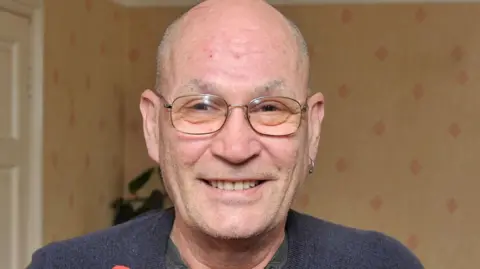 David Newton is smiling at the camera in an image from 2016. He is bald, has some stubble and wears thin framed glasses. He is wearing a navy jumper, with a grey top underneath and has an earring in his left ear. He is stood inside a room that has been papered with rust colour wallpaper that has red diamond shaped patterns. There is a white door behind him to his left and a green plant. 