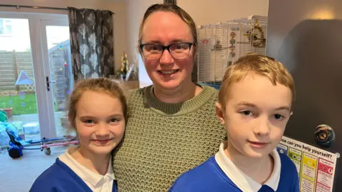 Alex Osborne/ BBC Image shows mum of two, Debbie Brouard, dressed in a dark green jumper, with hair tied back and wearing glasses, hugging her two children, Riva-Mae and Kian, wearing their blue and white school uniform. They're standing in the kitchen, by the fridge, smiling, with bird cage and garden visible in the background.