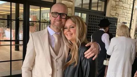 Nick and Maisie Ball at a wedding together. He is wearing a pale cream suit with  silk tie and pink shirt. He has his arm around Maisie, who is wearing a leather jacket and has long blonde hair. Behind them there are other wedding guests dressed formally. In the background there is a glass window where you can see the dining hall, set up with white tablecloths and pink decorations. 