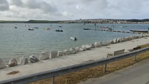Google The image shows a calm waterfront scene with a sheltered bay stretching across the frame. Several small boats float on the water, including motorboats, dinghies and a few sailing boats. The water appears steady with gentle ripples.