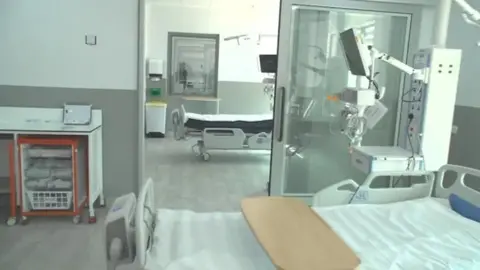 Hospital beds in individual rooms. The one nearest to the camera has a wooden tray across it. There is a monitor on a credenza beyond the bed. There is a table to the left with drawers of supplies beneath. There is another bed in the room beyond and an observation window beyond that.