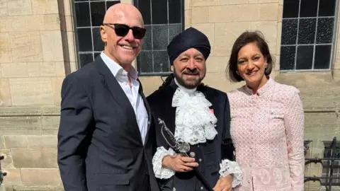 Pritpal Singh Nagi is wearing a black turban and navy blue trousers and jacket. The jacket has white frills on the chest and coming out of the sleeves, and has gold buttons. He is holding a slim sword. He is smiling and standing with a bald man with black sunglasses and a black suit jacket with a white shirt. There is a woman with a short brown bob wearing a pink and white patterned jecket