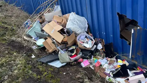 Anna Woodman Lots of household and personal belongings strewn across the green bank at the back of the blue tin unit. Amongst the stuff, we can see a PC screen, a handbag, brown boxes, some books and a photo frame. Can also see a big cut out square peering into the tin.