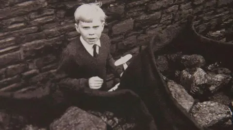 Getty Images A black and white image of an eight year old boy. His hair is white and he is holding a toy aeroplane, while sacks of coal are in the foreground.