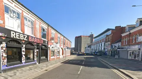 Google street view of Victoria Street South in Grimsby town centre. There are shops on either side most in two-storey red brick buildings. In the distance is a white building and a concrete tower block