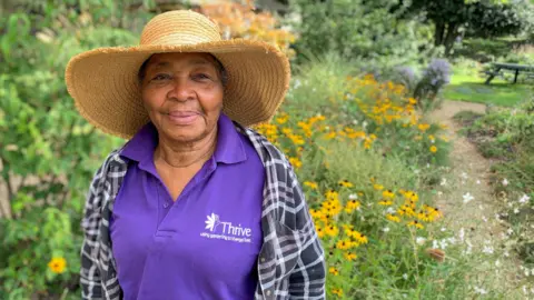 Thrive Cynthia, who is wearing a purple Thrive top and a brown hat is pictured standing in the garden. There are daffodils in the background and lots of greenery.
