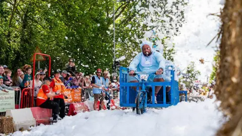 Dunmow Round Table Foam is strewn across the street with spectators watching on the side. There are trees in the background. A participant wearing a light blue poncho is cycling through it on his kart.