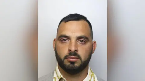 A mug shot of a man in his twenties, with brown eyes, a moustache and a beard. He is looking directly into the camera.