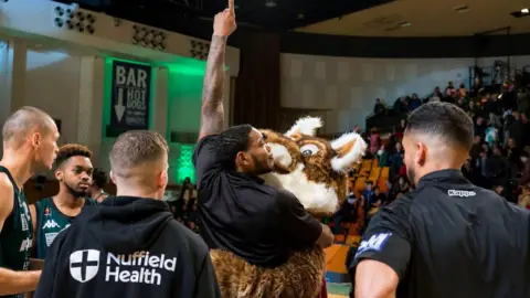 Plymouth Raiders Basketball Club Kinu Rochford holds an arm in the air as he hugs a mascot with other players standing around him.