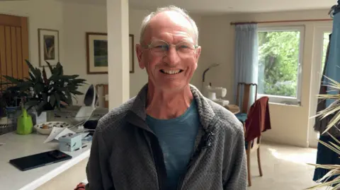 A white man in his 60s smiling at the camera. he has grey, set back hair and is wearing a blue T-shirt and grey fleece. He is in his home.