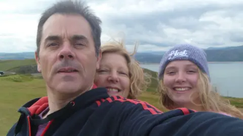 Rodina Obrázek muže, ženy a dospívající dívky se usmívá na kameru stojící na kopci. Hory a moře jsou na pozadí.