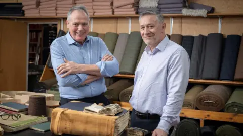 Kirsty Anderson/Lovat Mill Two grey-haired men in a room surrounded by bales of tweed fabric. A large, old book is on a desk in front of them. 