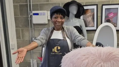 Rose Collins A woman wearing an apron with Rose Collins written on it. She has her arms spread out showing ostrich feather hats in front of her. There is a mannequin behind her wearing a black hat and pictures of women wearing hats.