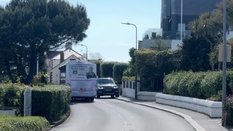 A road with a hedge on the left and a pavement and hedges on the right. There is a white bus passing a black car and the car in opposite directions. The car is coming towards the camera and two of its wheels are on the pavement.