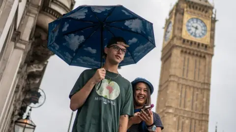Two tourists under an umbrella in central London outside Big Ben. The young man is wearing a baseball cap, t shirt and glasses. The lady next to him is wearing a hat and smiling while holding her mobile phone.