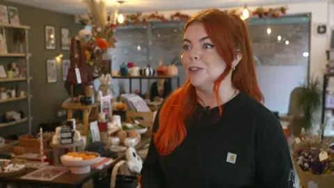 BBC Charlotte Brennan with long, bright orange hair and a black sweater stands in front of her gift shop items which includes homeware,ghost-shaped pillows, dried flowers, candles and ornaments