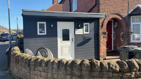 The shed has a door and has a flat roof. It is painted black and sits next to the front door of the house. 
