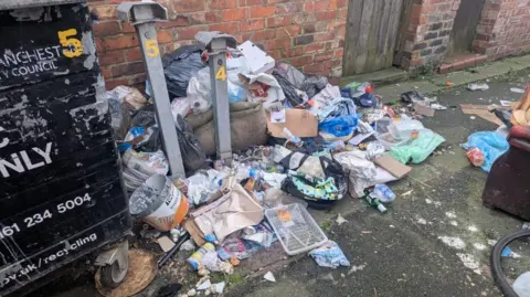 Rubbish strewn across the floor by a large, black communal bin, in an alleyway 