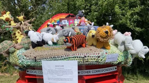 A colourful knitted display of Noah's Ark complete with animals. The knitted postbox topper is on top of a red postbox. There is a rainbow and lots of knitted animals. The sun is shining. 