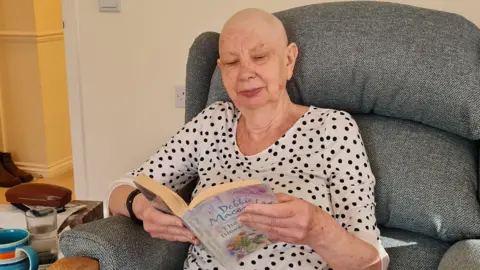 Diana Brookes sitting reading a book in a white and black polka dot t-shirt, she is sitting in a grey armchair.