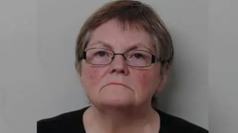 Police mug shot of Jay Reid - a woman dark, greying hair and glasses, looking unhappily into the camera