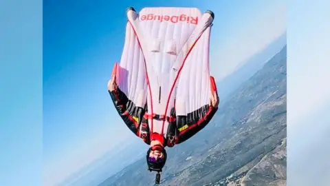 Byrne family Man - Liam Byrne - wearing wingsuit, upside down in mid air and smiling at camera.