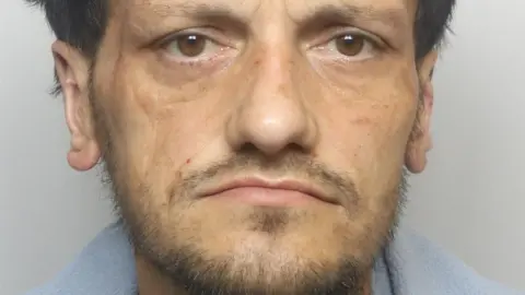 A man with a dark beard stares into the camera in a police custody shot.
