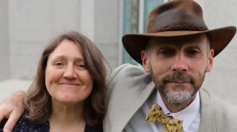 Family handout A man with his arm around a woman's shoulders. They are both smiling at the camera. The man is wearing a brown leather hat and a light coloured suit with a yellow bow tie. The woman has mid-length brown hair.