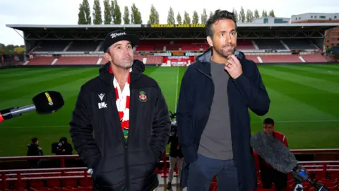 PA Media Rob Mac and Ryan Reynolds are pictured in the Wrexham Racecourse stadium with the football pitch behind them. They have contemplative expressions and two broadcast microphones can be seen pointed at them.