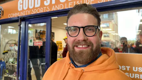 Andrew Turner/BBC Jack Jay standing outside a shop with an orange and blue shop front. He is standing to the right of the frame looking directly at the camera and smiling. He has a trimmed fair beard and short swept-back hair and is wearing an orange hoodie and a pair of black glasses.