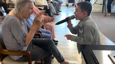 ITV News West Country Lucciano singing on his knees with a microphone before an elderly care home resident