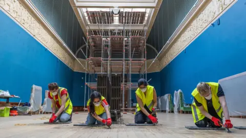 Victoria Art Gallery Conservationists working on the floor of the gallery wearing hi-vis jackets and facemasks. They are all in a line kneeling on the floor with scaffolding in the background.