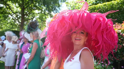 A woman, stood next to friends in the background, is wearing a very bright pink hat with long, thick tasles drooping from it.