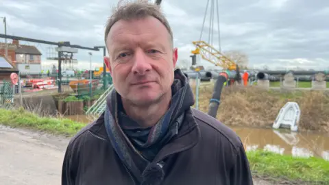 Ian Withers is standing next to a pumping station by a river. He is wearing a black fleece and a scarf.